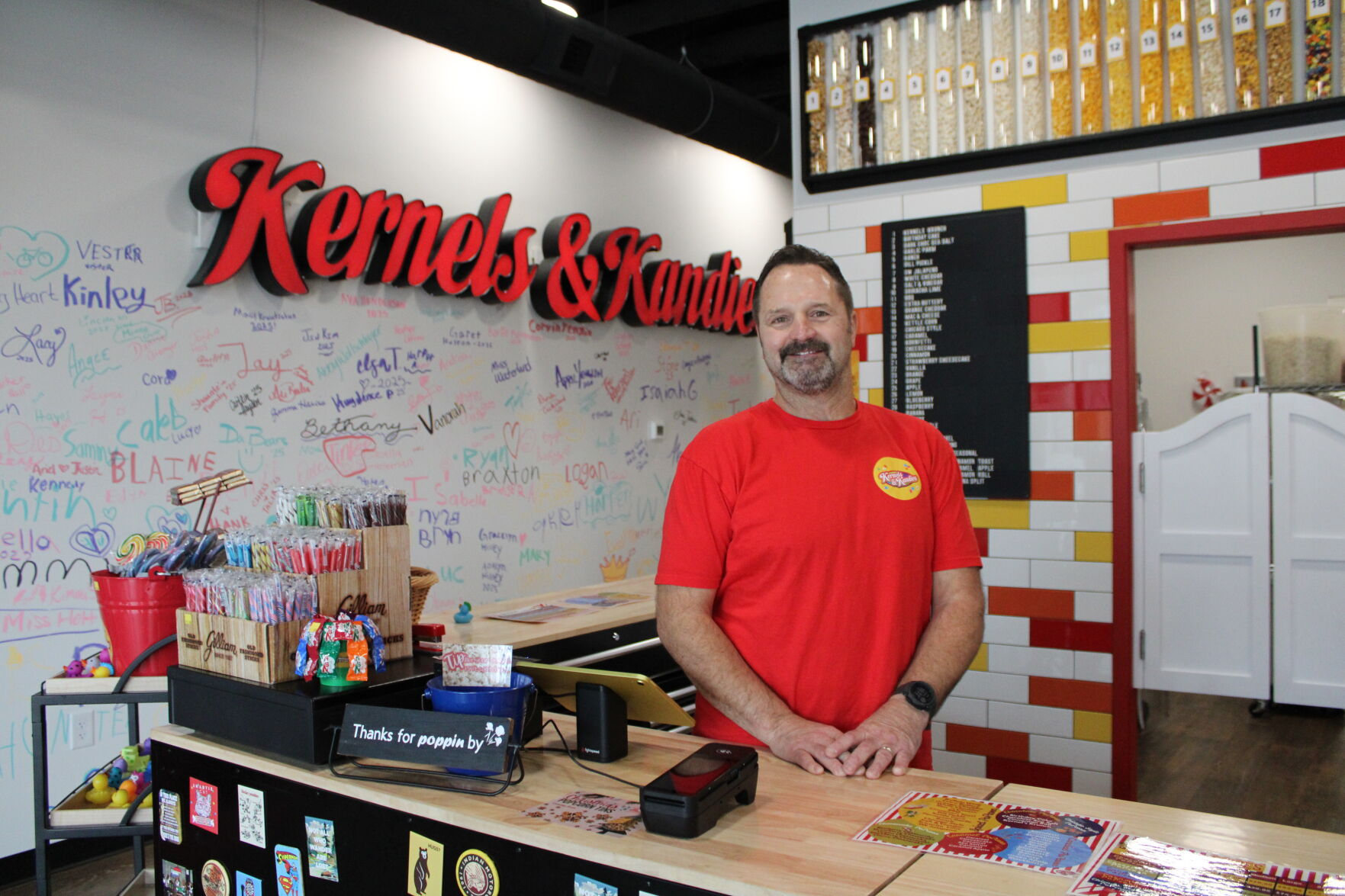 Owner behind the Kernels & Kandies counter with the popcorn bins and signature wall